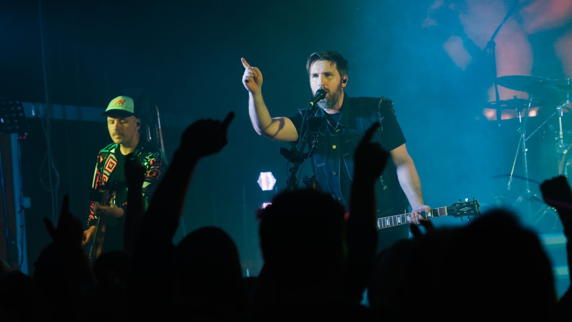 Band performing on stage with blue lights and screen behind them; crowd in front.