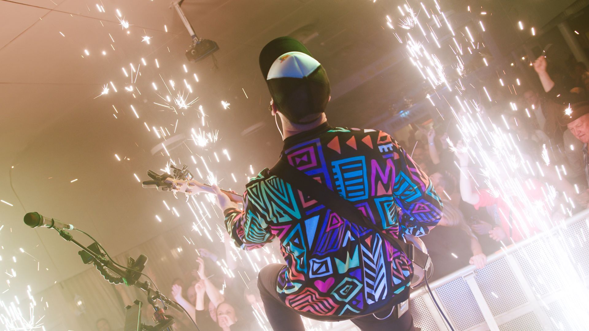 Person playing guitar on stage with fireworks. Brightly colored shirt and a crowd in the background.
