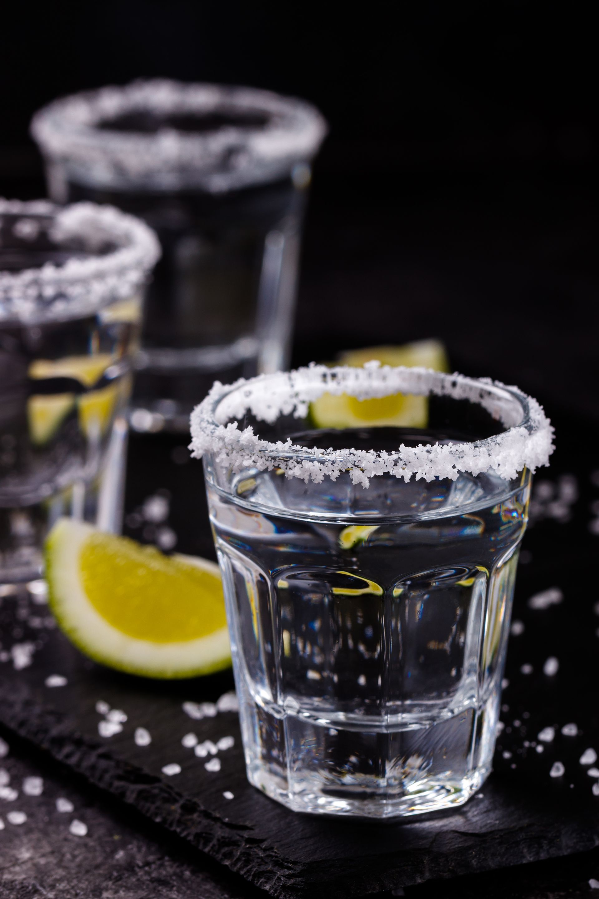 Three tequila shots with salt rims, and lime slices on a black surface.