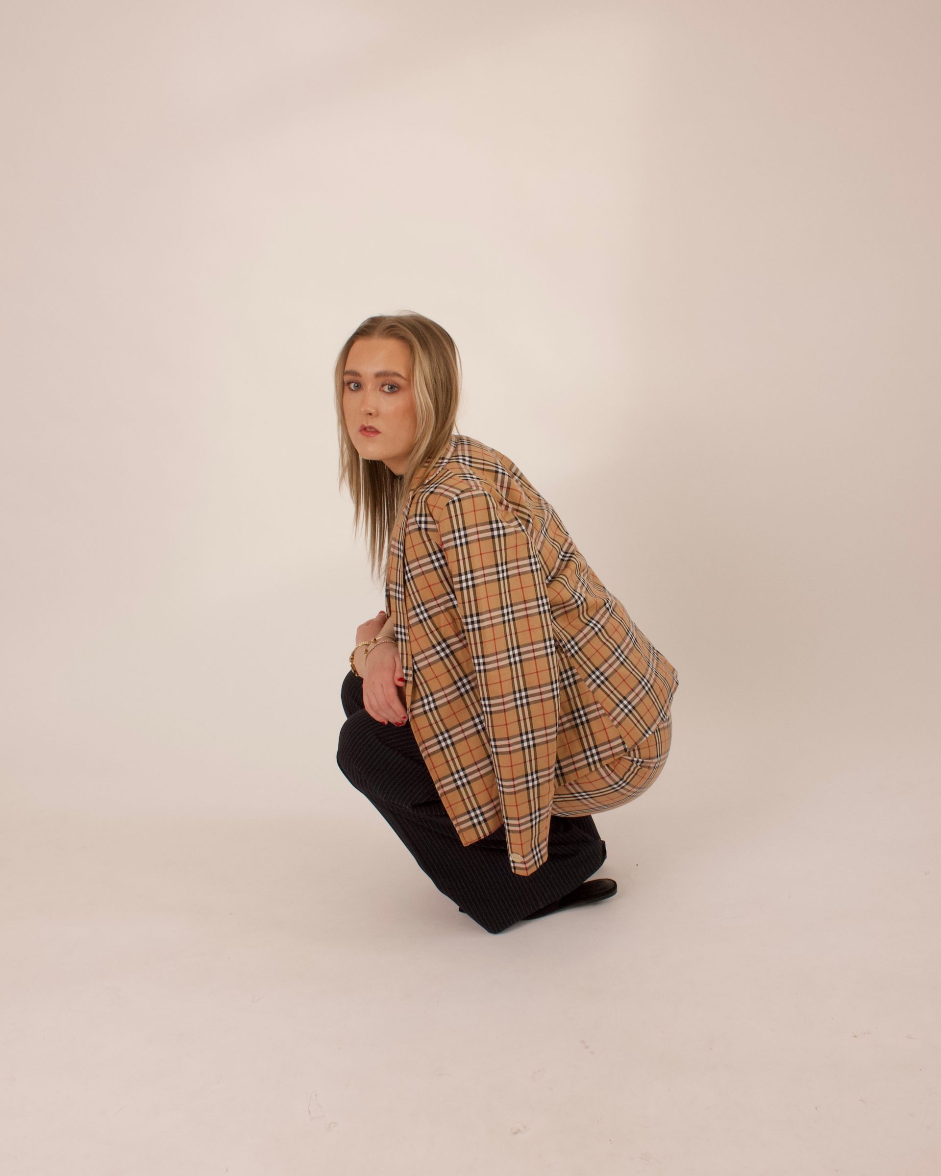 Woman crouched, wearing plaid jacket and black pants, against a plain background.