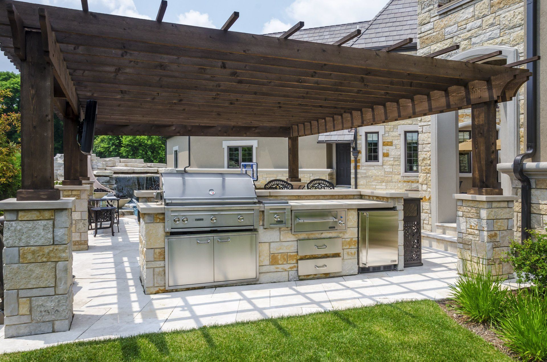 An outdoor kitchen with a grill under a pergola in the backyard of a house.