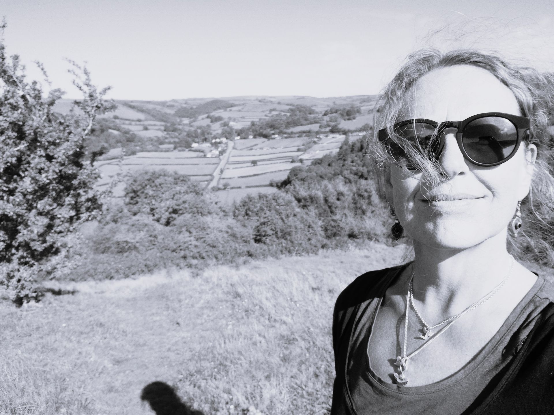Woman with sunglasses smiles at the camera, overlooking a valley. Black and white photo.