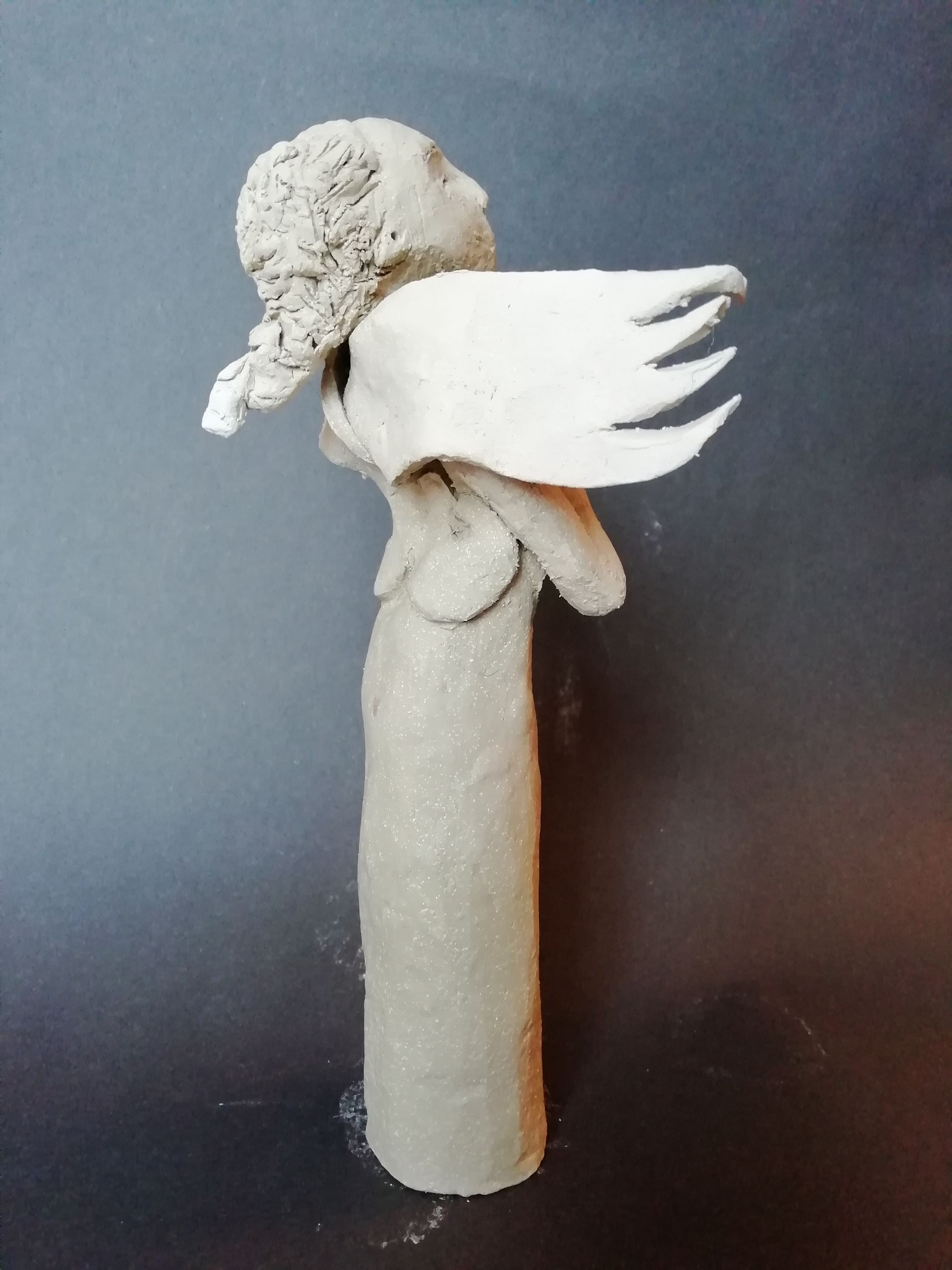 Clay sculpture of an angel with folded wings, looking upward.