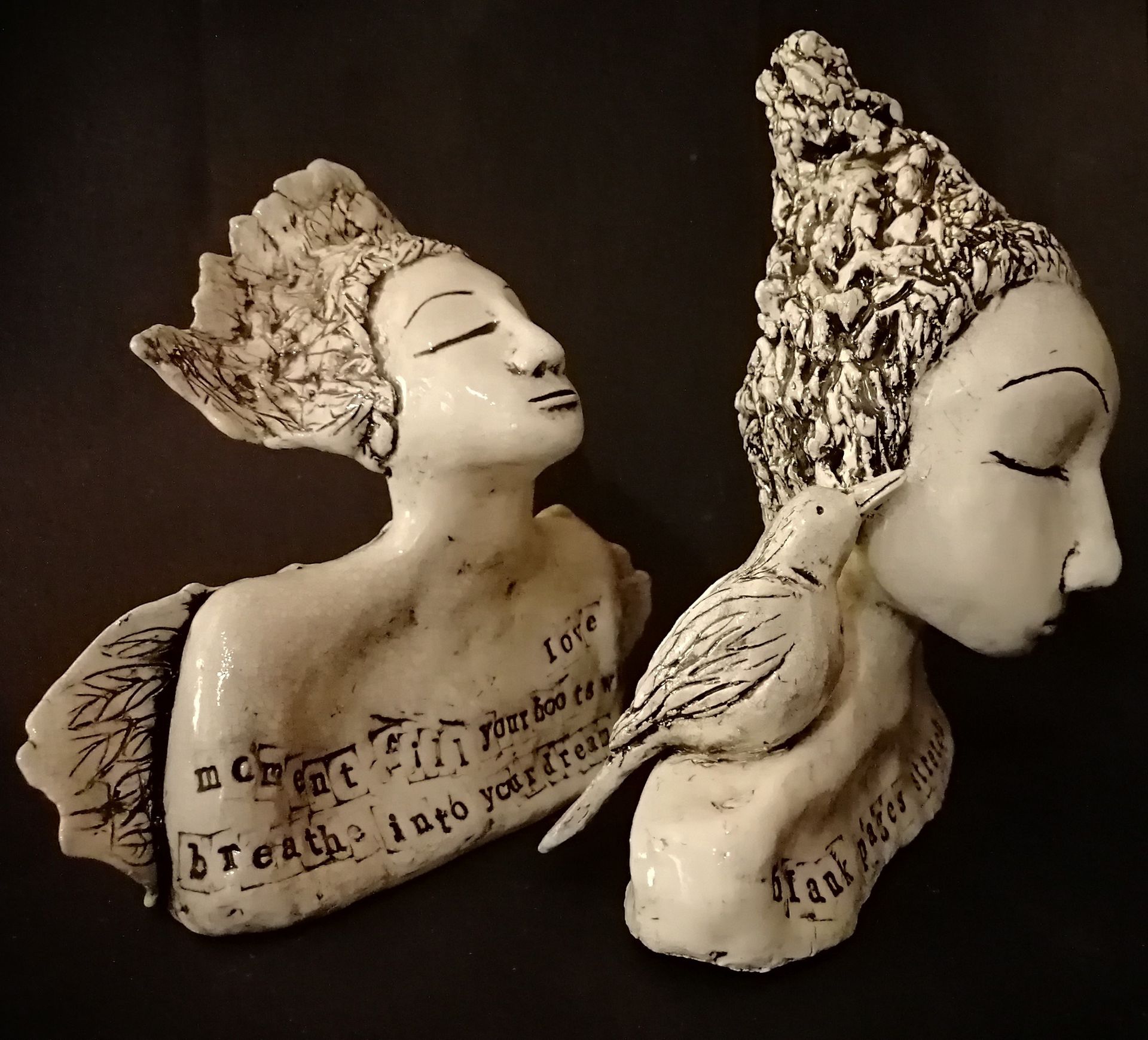 Two white ceramic sculptures of serene female busts with closed eyes, one with a bird, text inscribed.