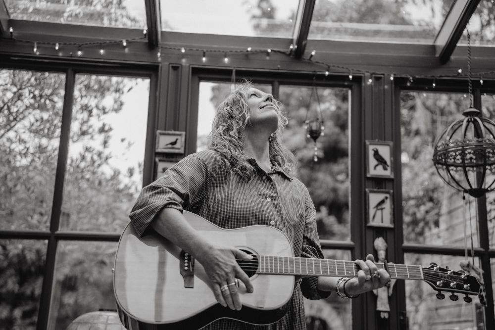 Woman playing acoustic guitar in a sunroom, looking upwards, surrounded by windows and string lights.