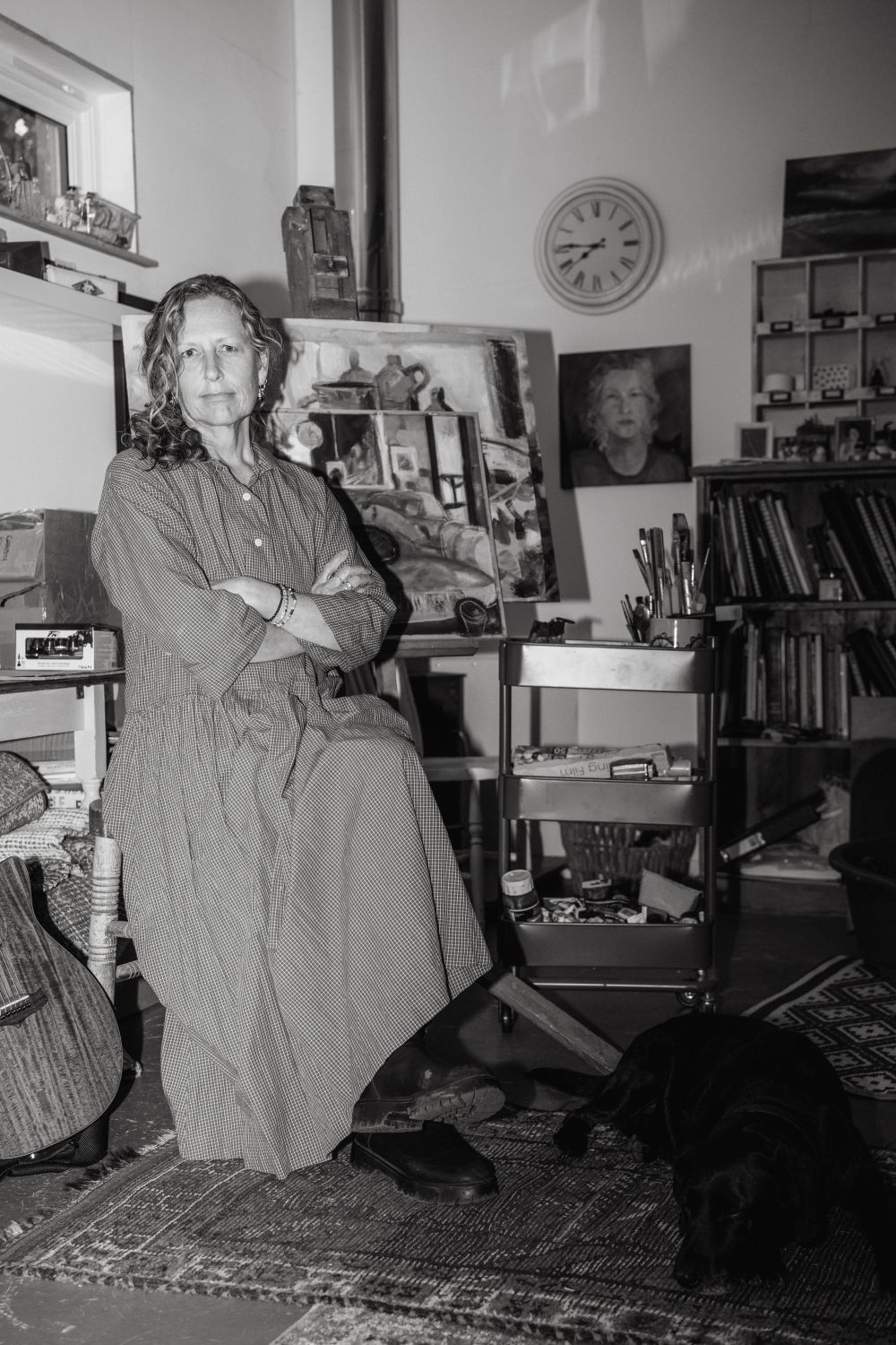 Woman in a dress sits with arms crossed, in a room with paintings, clock, and a dog.