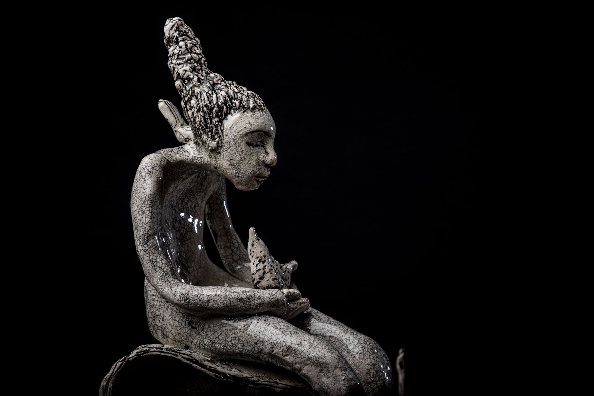 Ceramic sculpture of a seated figure, holding a small star, with eyes closed, against a black background.