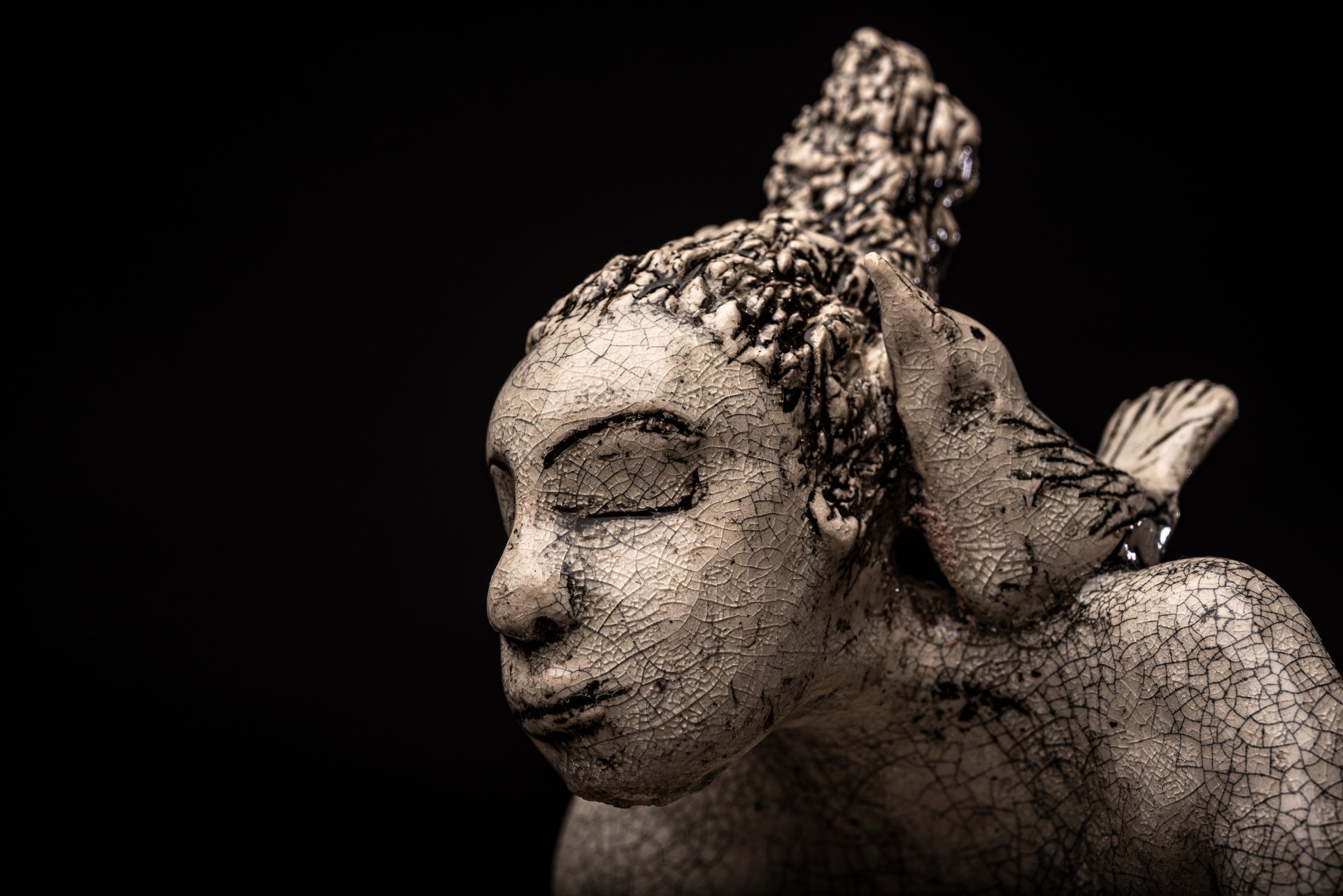 Ceramic bust of a woman with closed eyes, bird perched on her shoulder, against a black background.