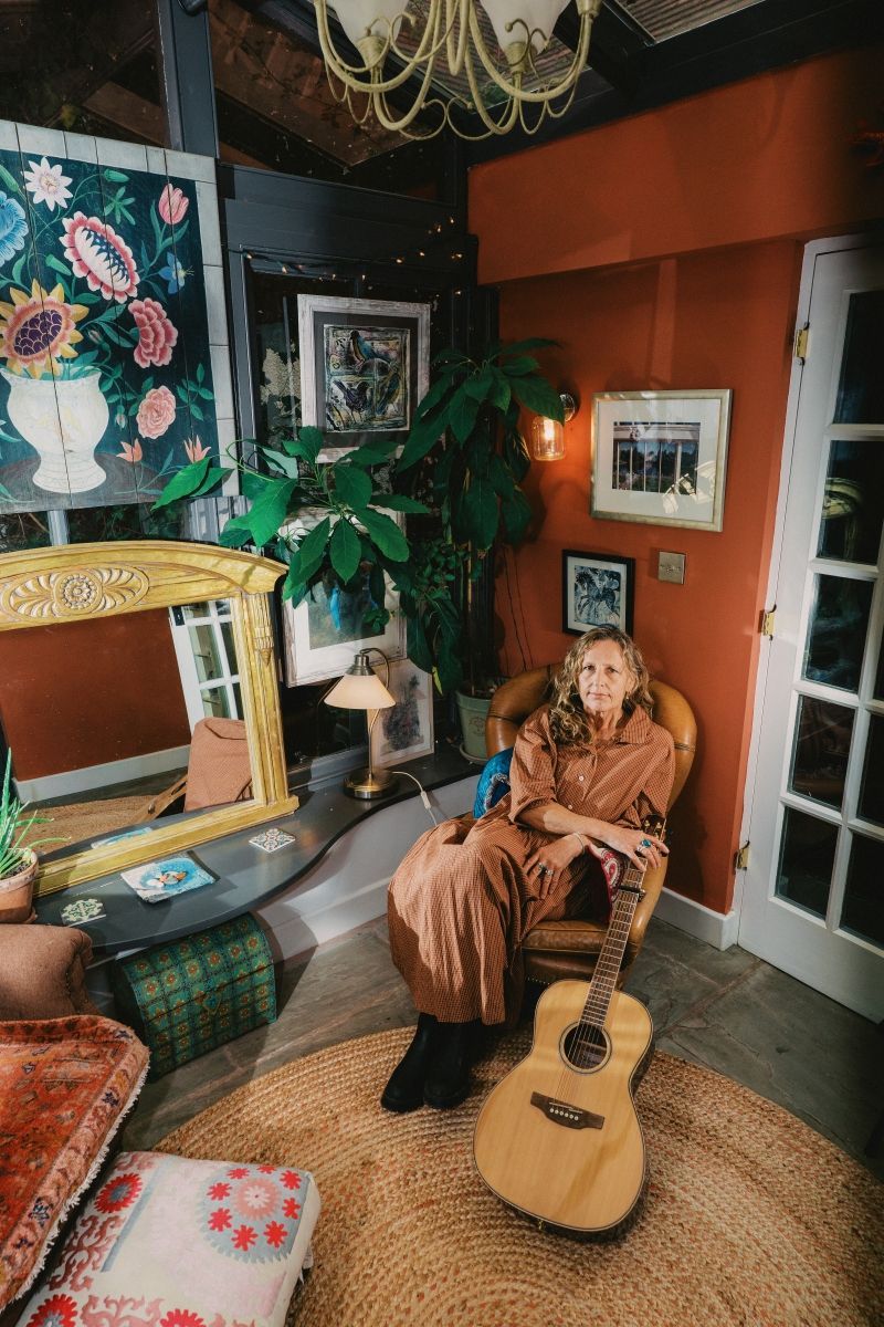 Woman in brown jumpsuit seated with acoustic guitar, near painting, mirror, and plants. Interior with warm colors.