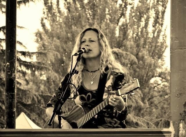 Woman with long, wavy hair sings and plays guitar on an outdoor stage. Sepia-toned photo.