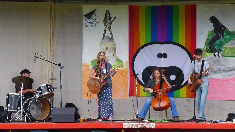 Band performing on stage with drums, cello, guitars, colorful backdrop.