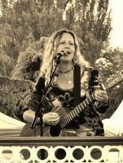 Woman with long hair playing guitar and singing into a microphone outdoors.