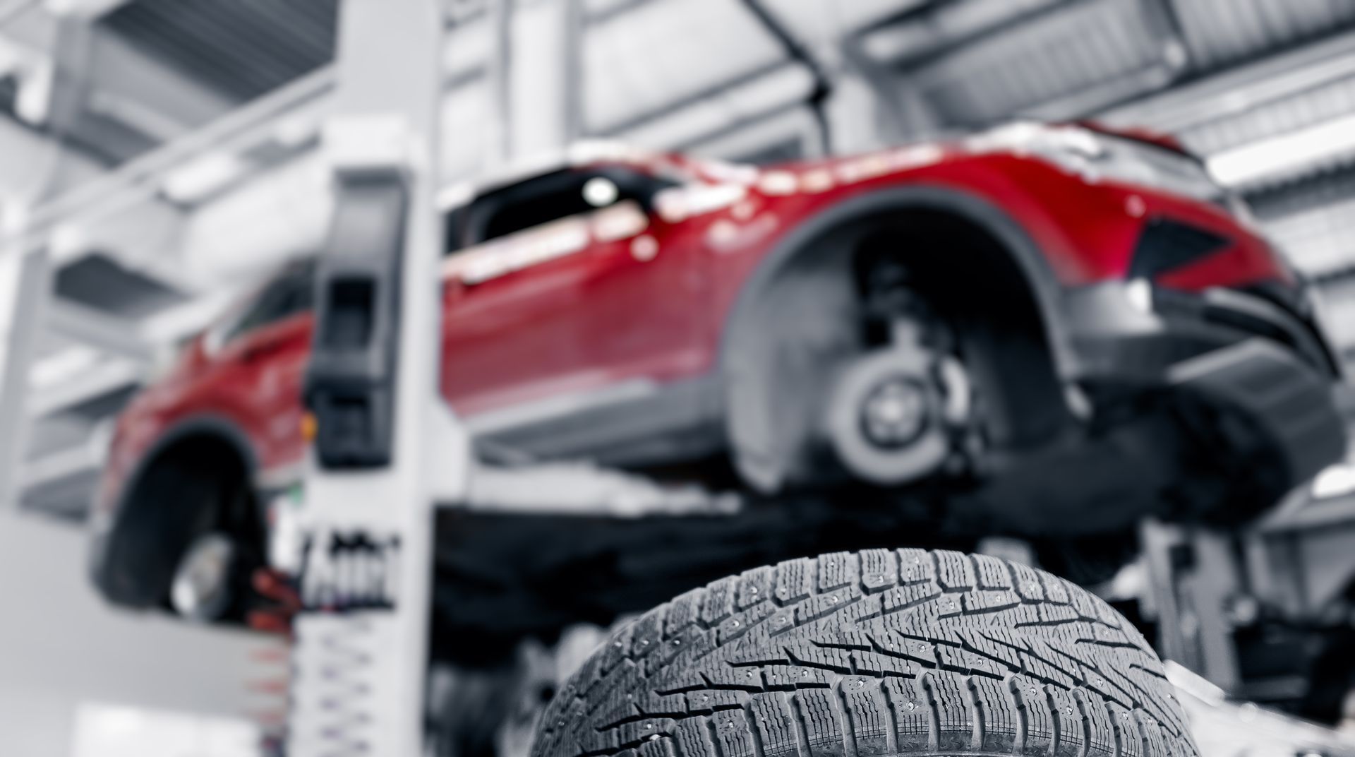A red car elevated on a mechanic's lift in a garage, with a removed tire in the foreground.