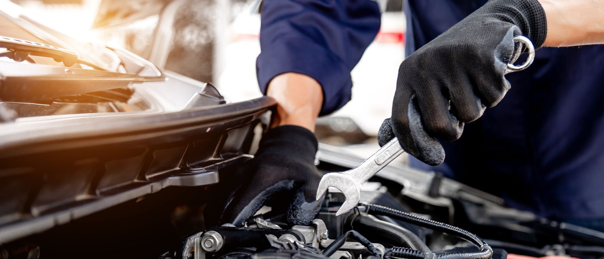 Mechanic in dark blue uniform and black gloves using a wrench to repair a car engine under the hood.