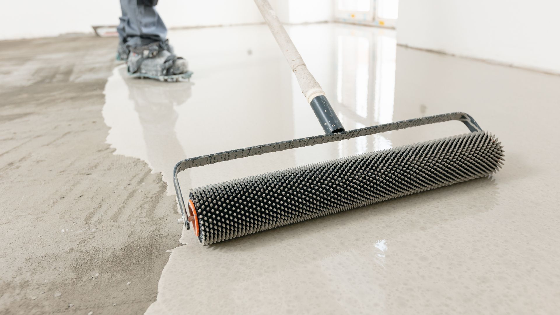 A person is using a roller to spread concrete on a floor.
