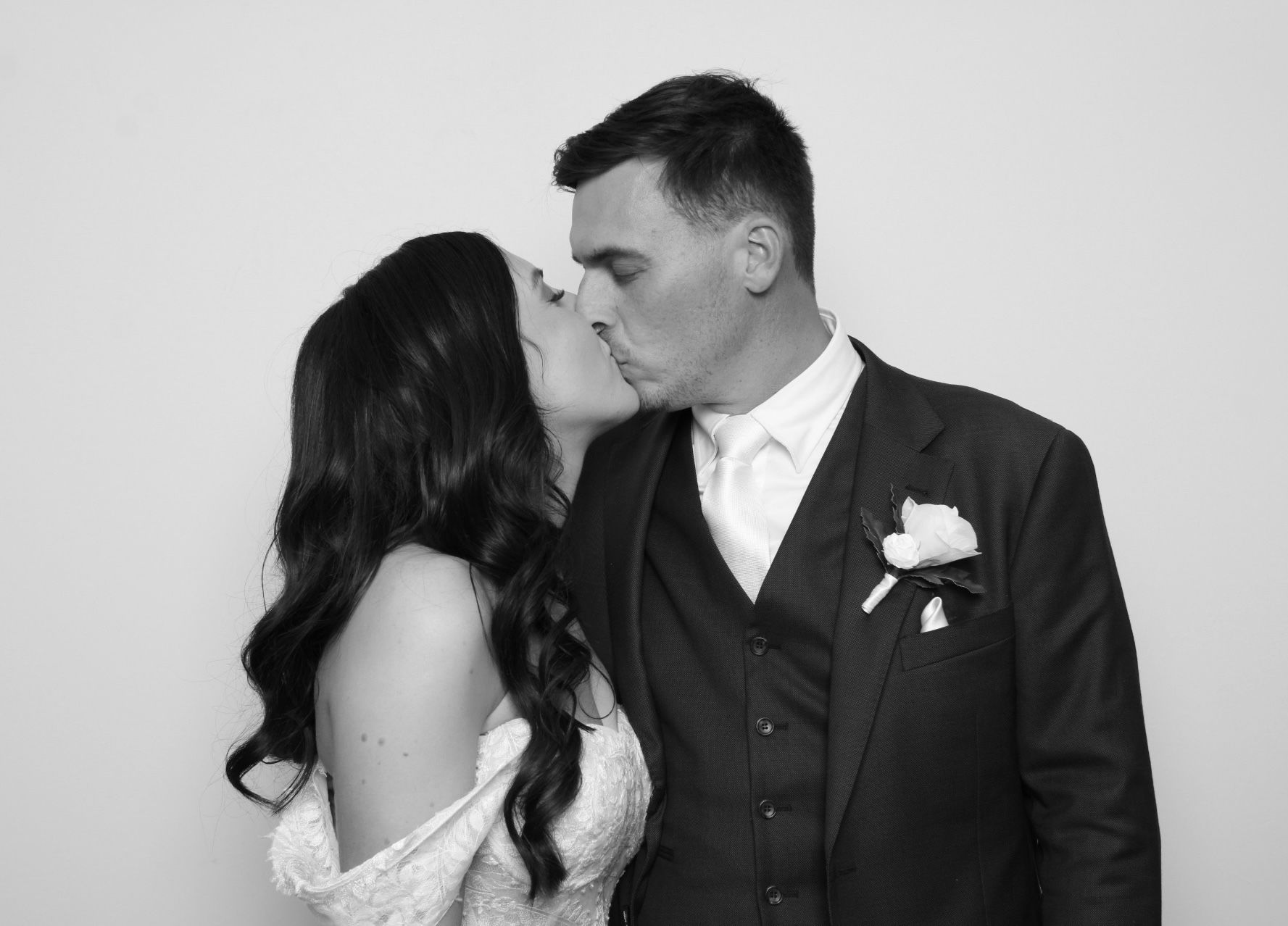 Couple kissing, bride wearing a white dress, groom in a dark suit, photo booth setting.