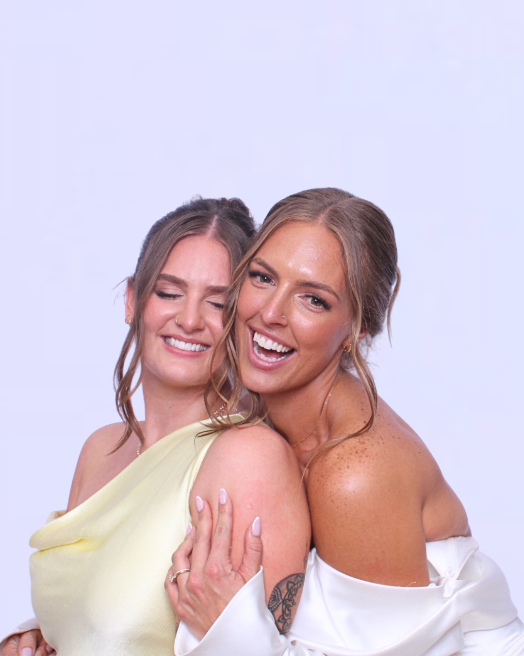 Photo Booth. Two women embrace, smiling. One in a yellow dress, the other in white, against a white backdrop.