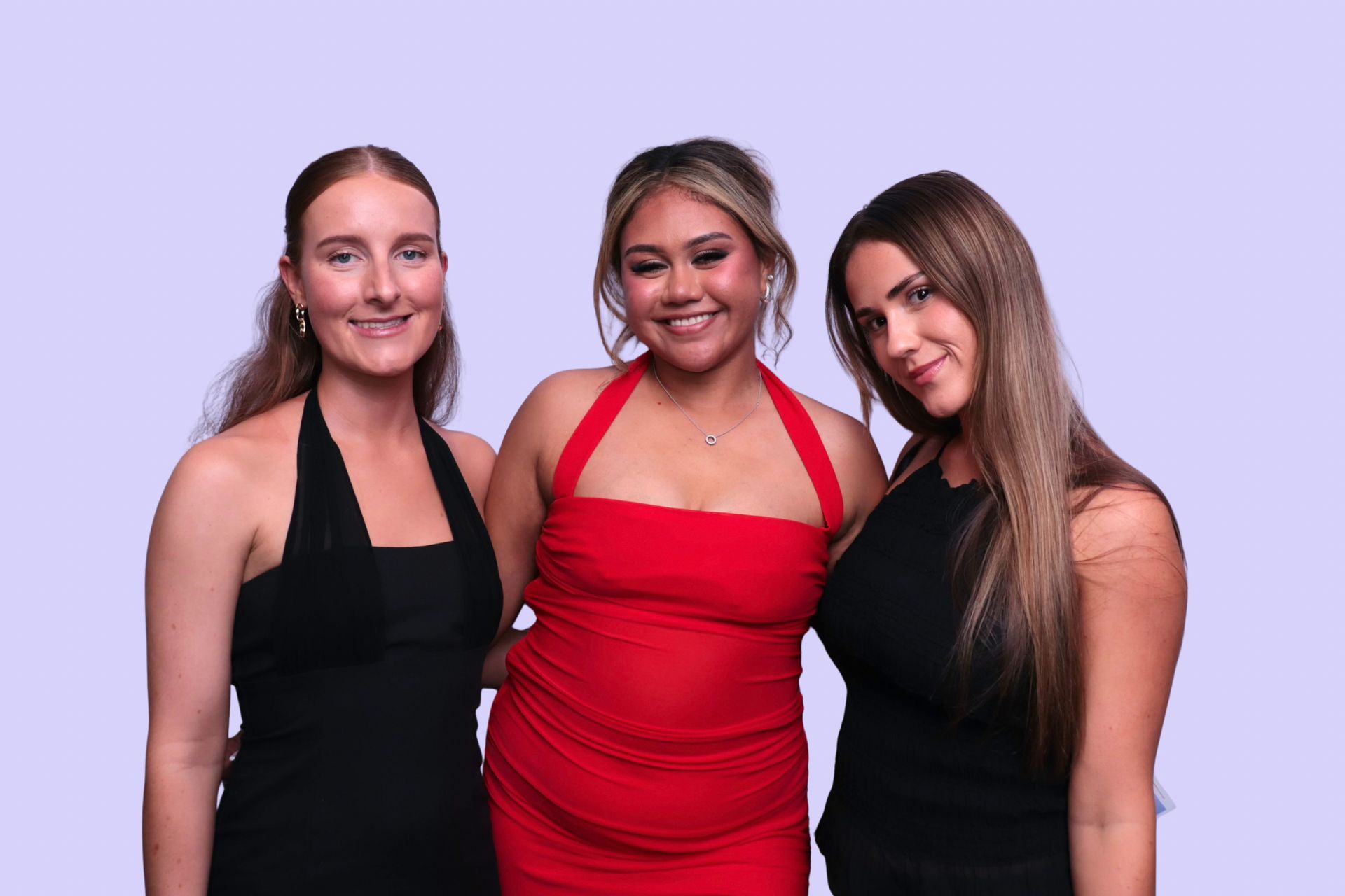 Three women in formal dresses pose, smiling in birthday Photo Booth. One in red, two in black, against a plain background. 21st birthday party. 