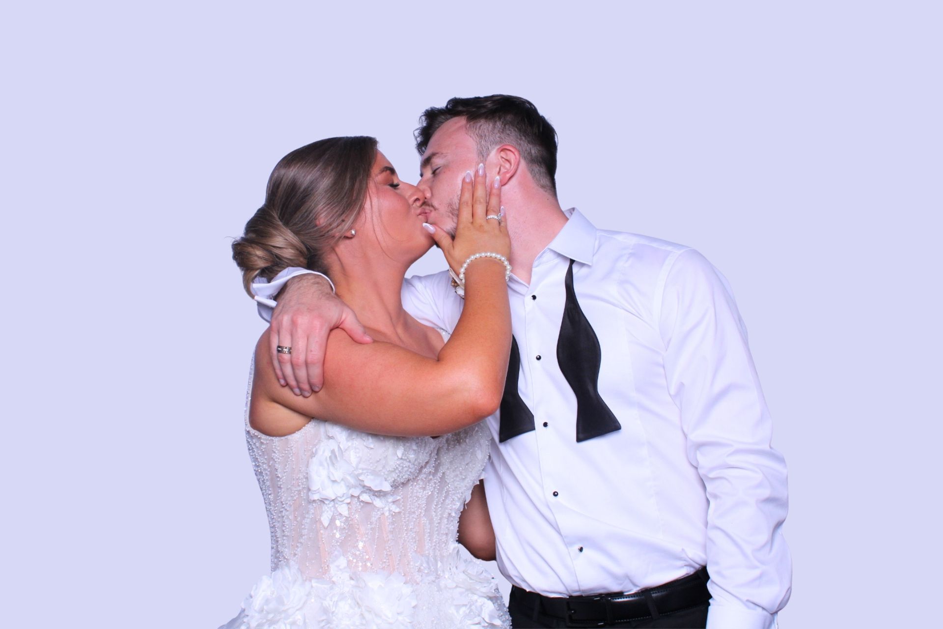 Wedding couple kissing; bride in white gown, groom in tuxedo, against light purple background. Photo Booth wedding