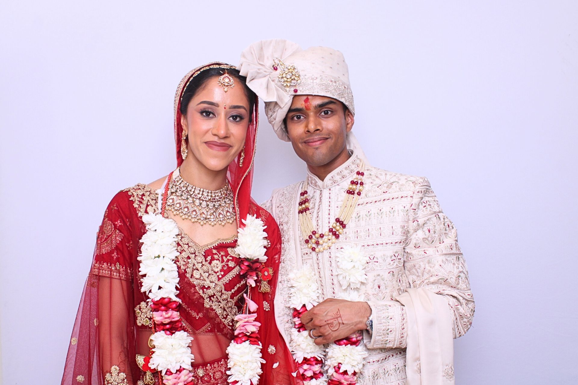 Bride and groom in traditional Indian wedding attire, smiling, with garlands, against a plain background in Photo Booth. 