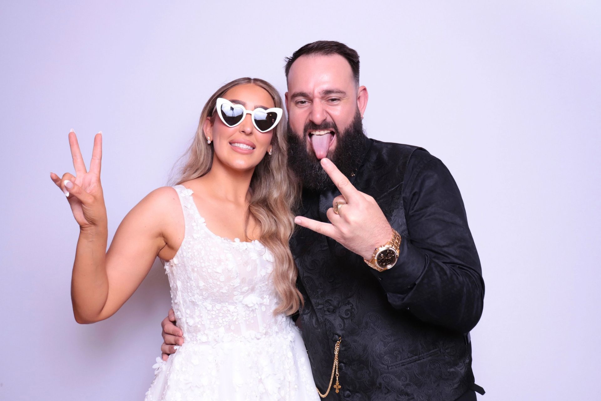 Couple posing, woman in white dress with heart-shaped glasses, man with beard, making rock hand gesture.