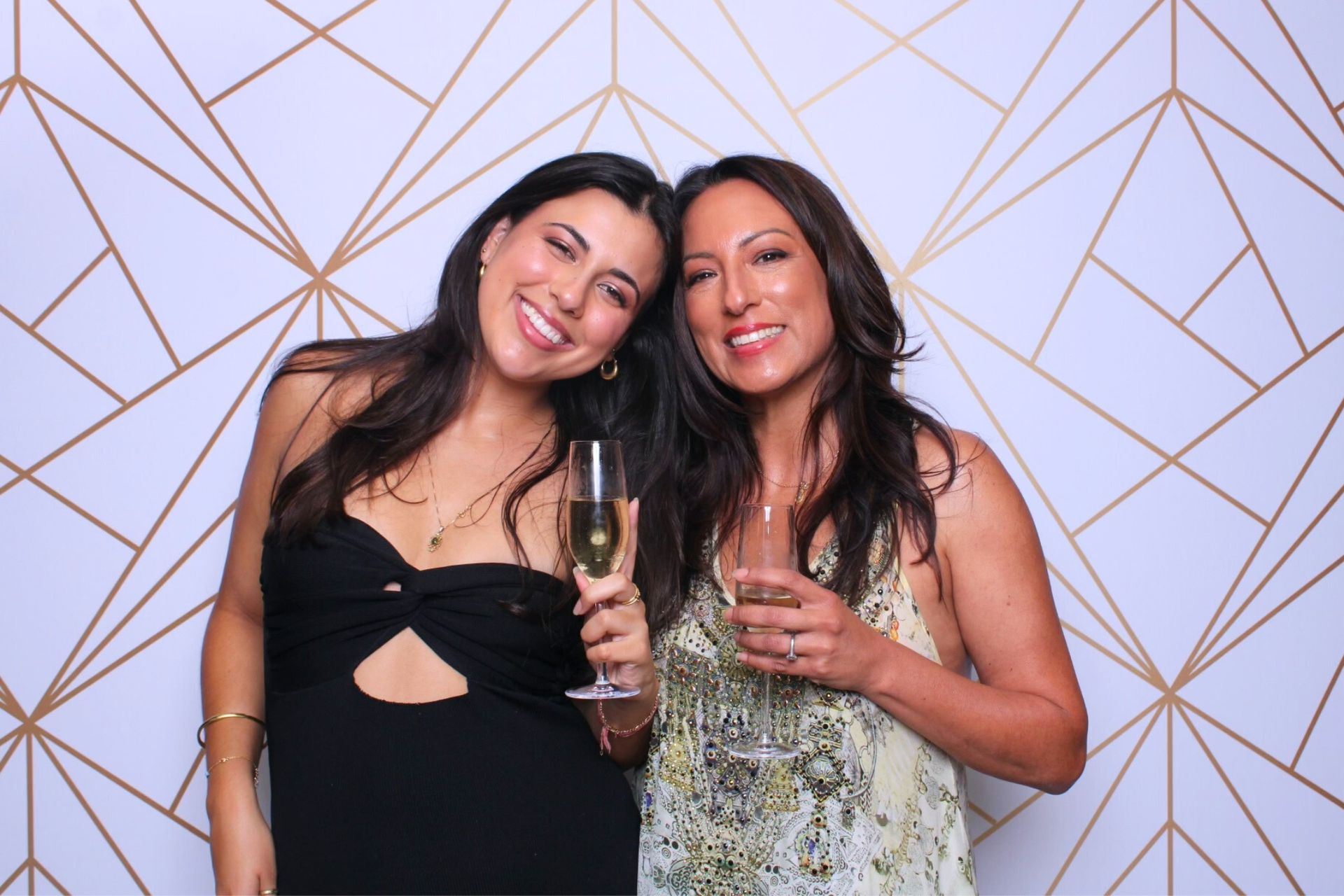 Two women smiling in Photo Booth, holding champagne glasses in front of a geometric gold and white background.