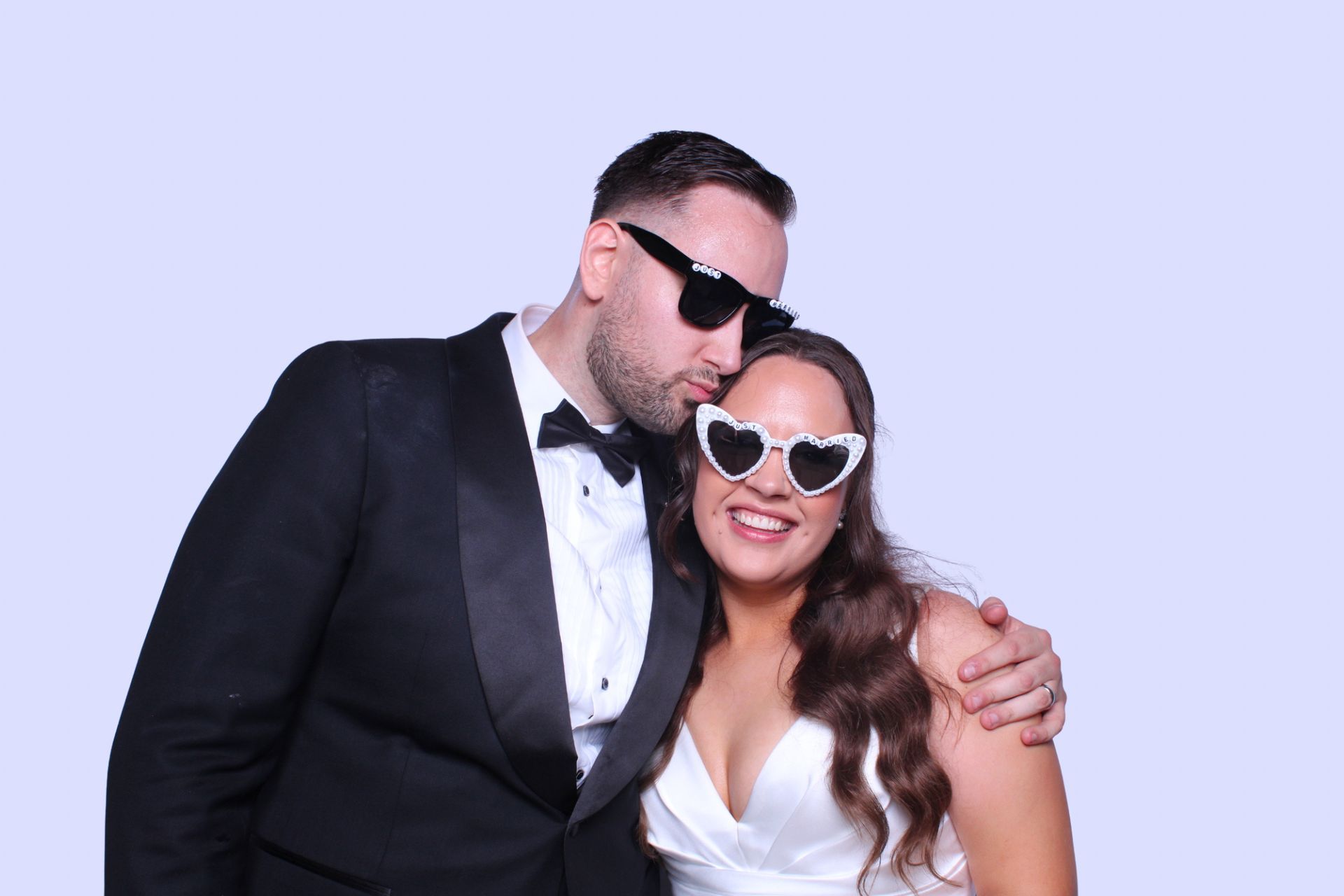 Photo booth couple wearing sunglasses embrace, man kisses woman's cheek, against a light blue backdrop.