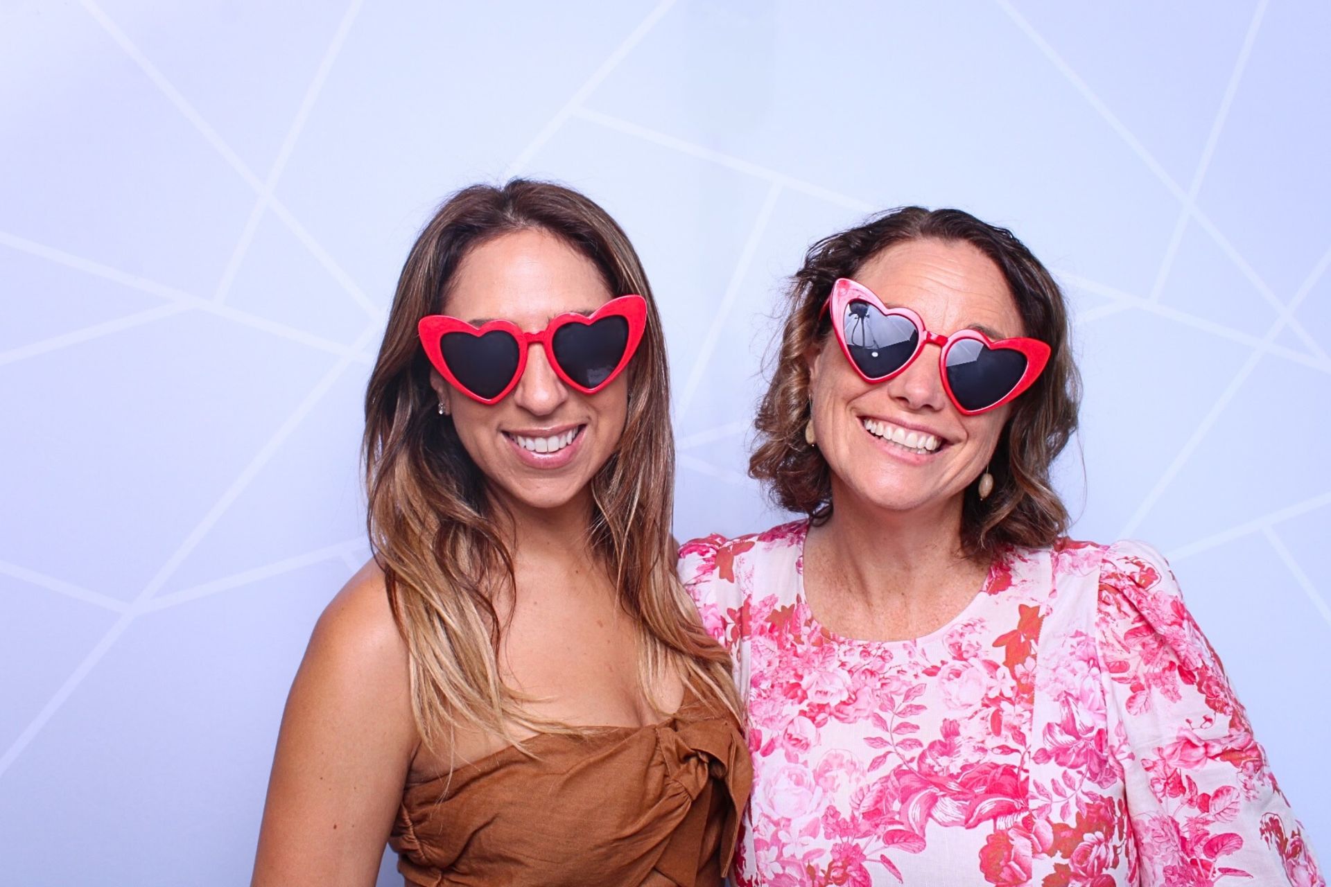 Two women smiling, wearing heart-shaped sunglasses, posing in front of a geometric backdrop PHOTO BOOTH