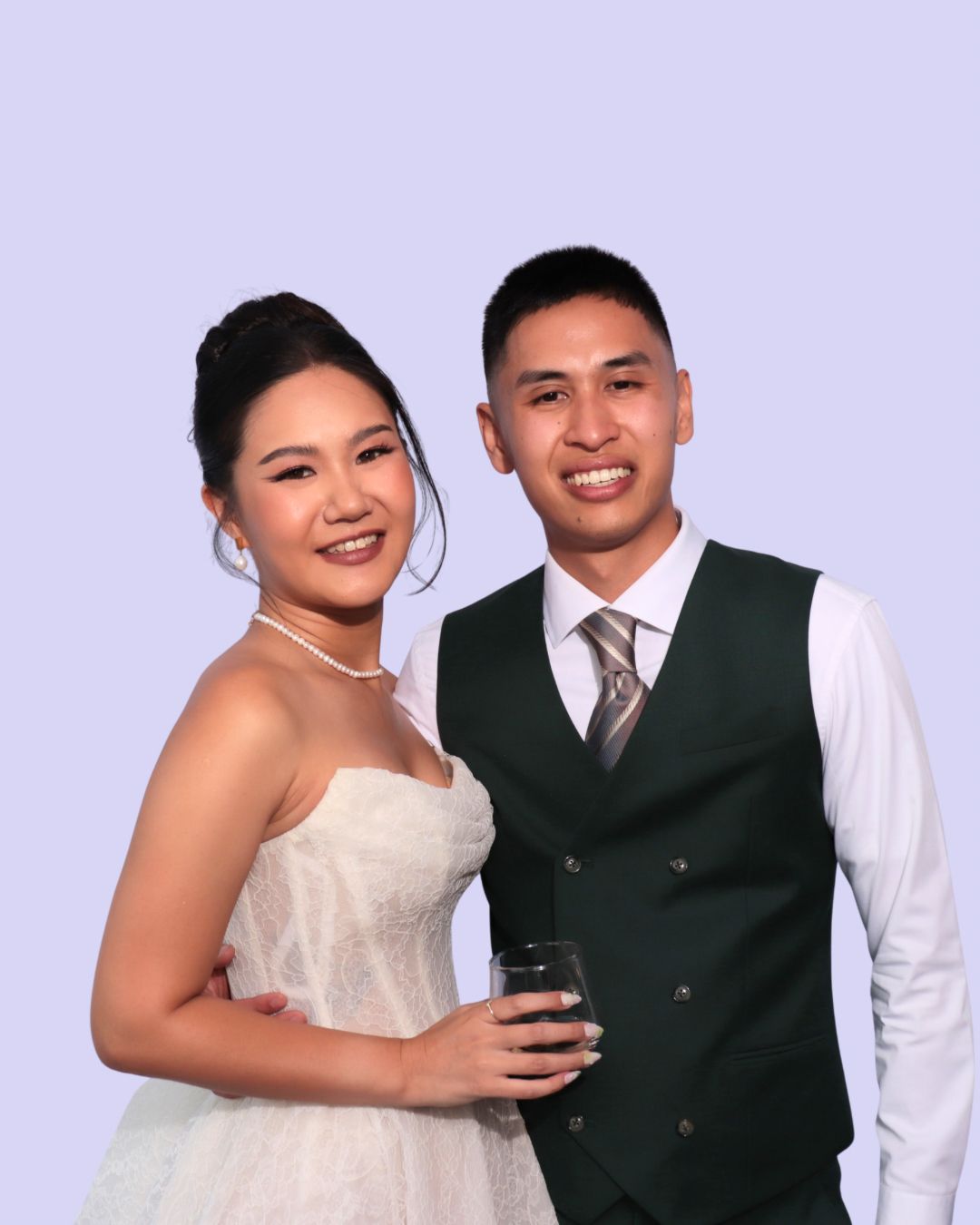 Couple smiling, embracing. Woman in white dress, pearls; man in green vest, tie, holding glass. Wedding photo booth