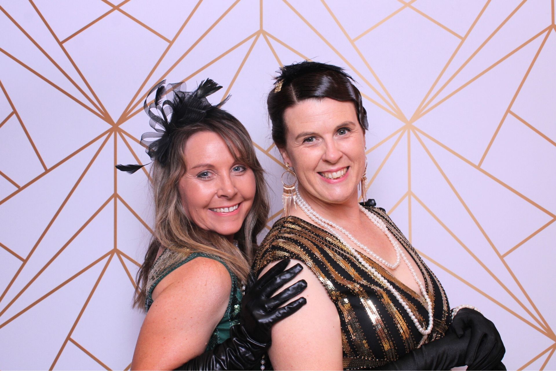 Two women smiling at a photo booth; art deco backdrop, flapper attire.