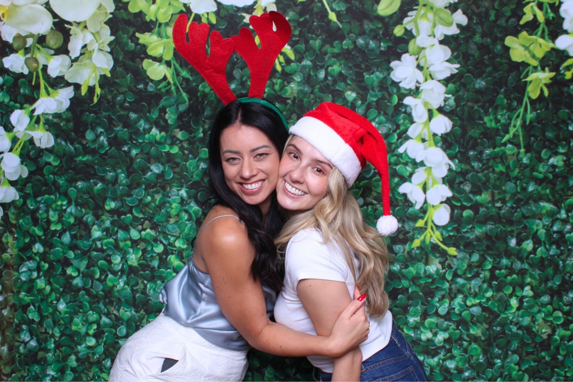 Two women smiling and hugging in front of a green hedge backdrop. One wears reindeer antlers, the other a Santa hat.