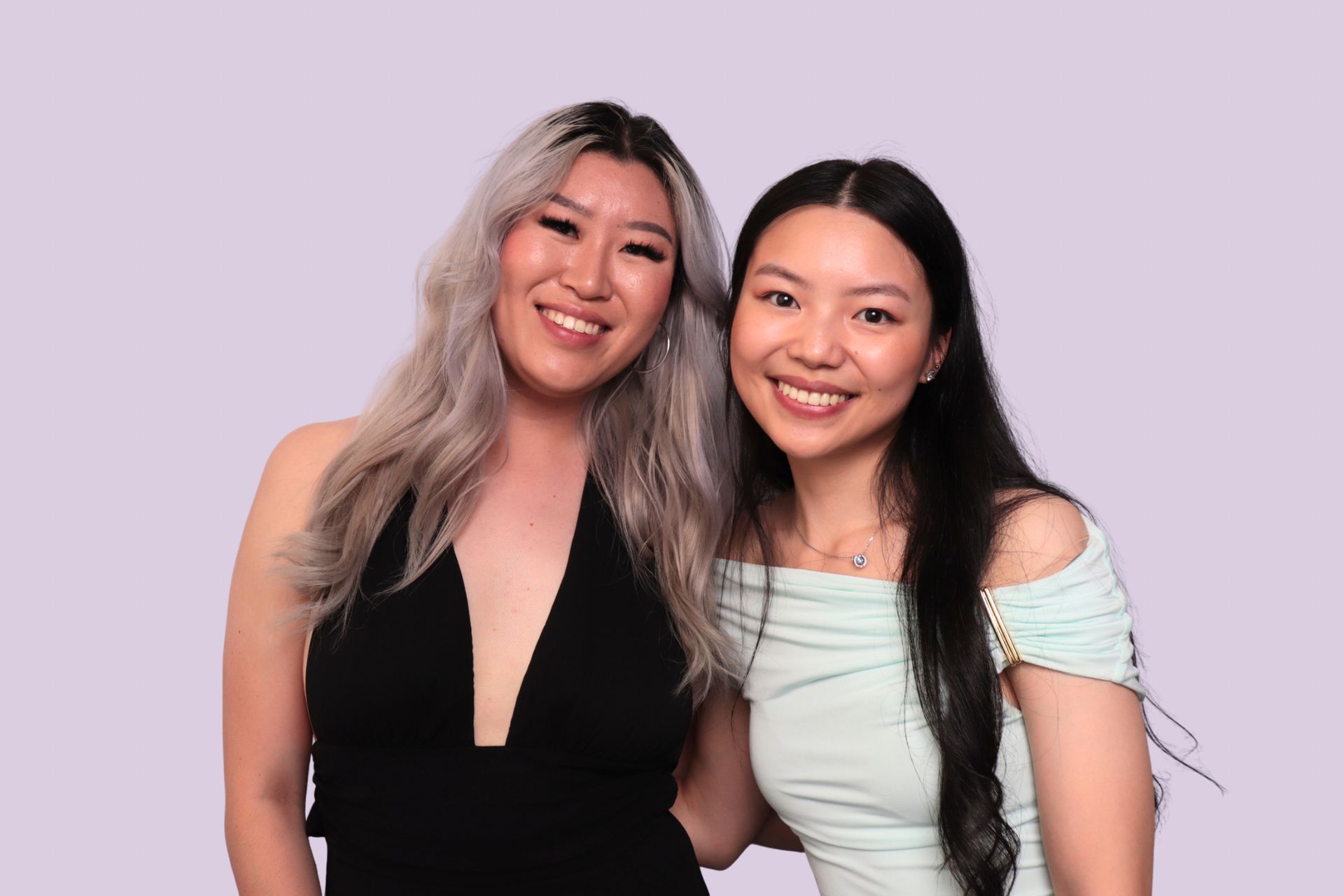 Photo Booth. Two smiling people pose against a lavender background. One wears a black dress, the other a light blue top.