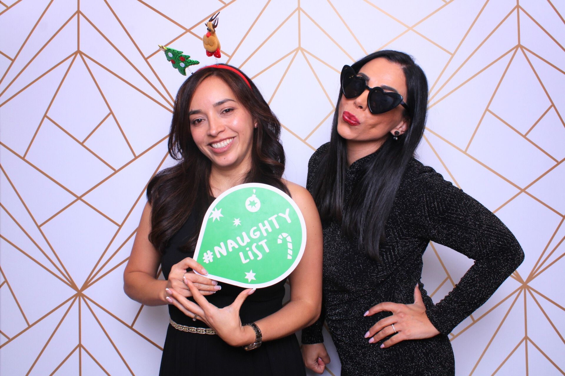Two women pose in front of a geometric backdrop. One holds a 