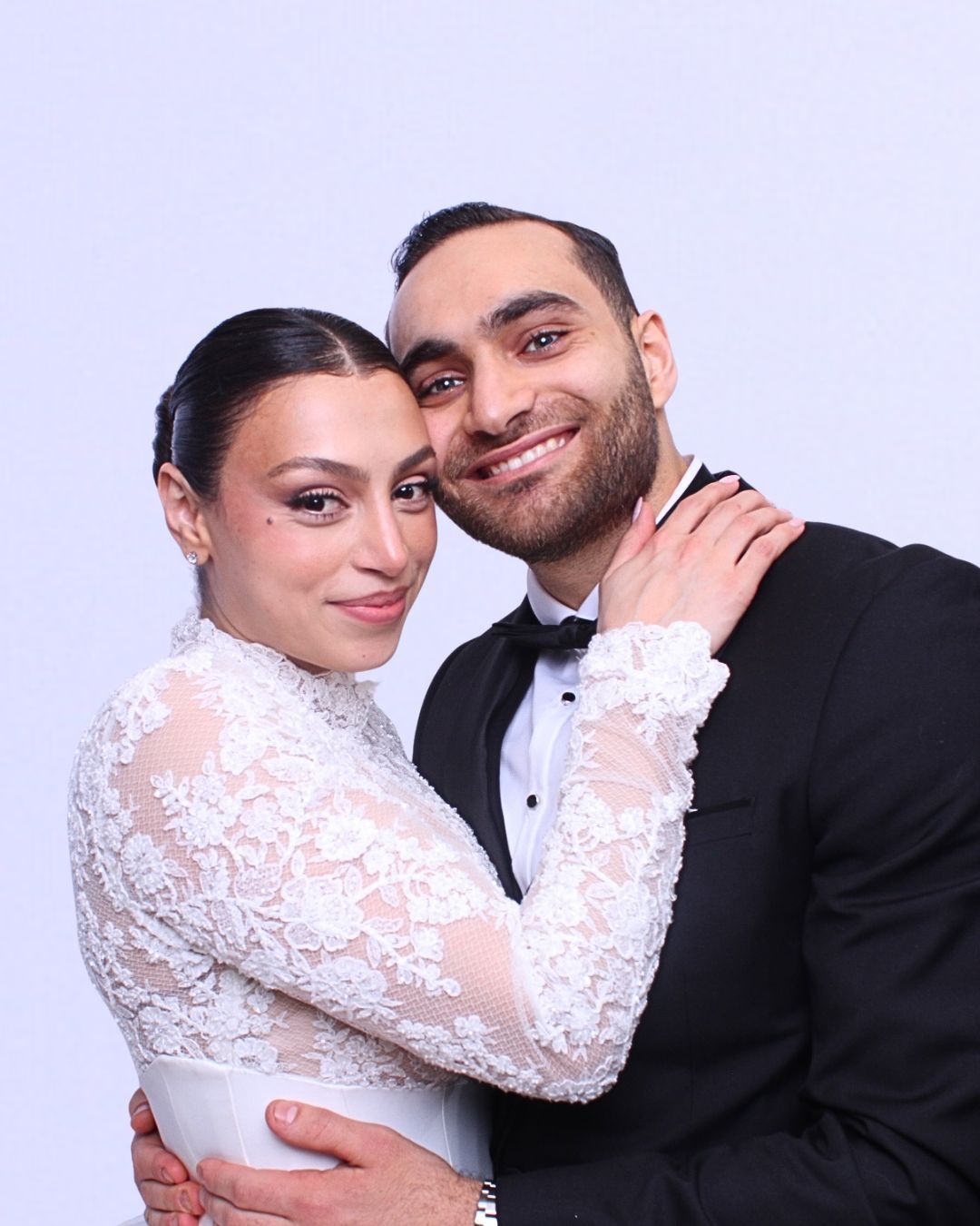 Bride and groom pose closely in Photo Booth. The woman in a white lace dress smiles, man in a tuxedo smiles, cheek to cheek.