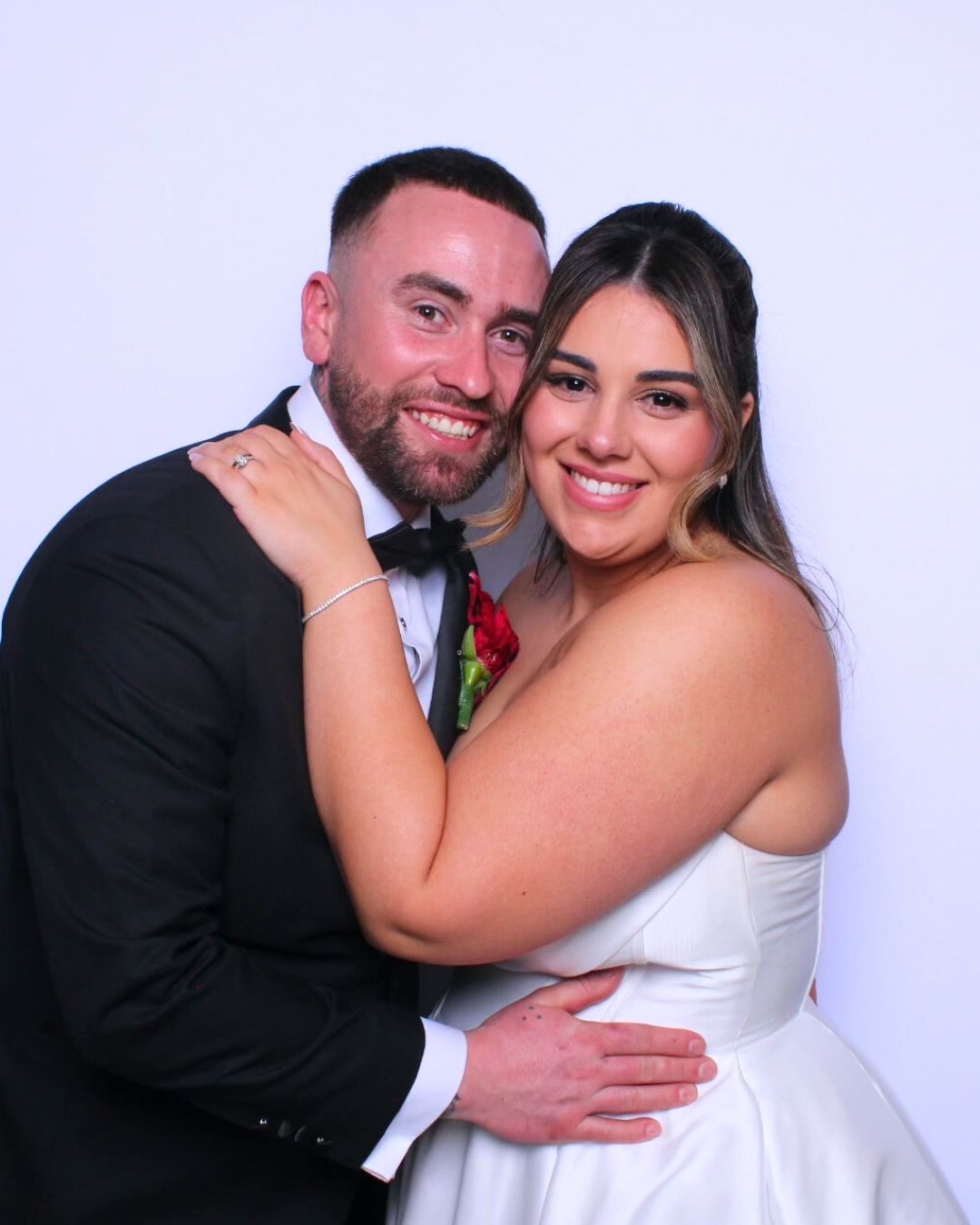 Newlyweds smiling in a photo booth. The bride wears a white dress and the groom a black tuxedo.