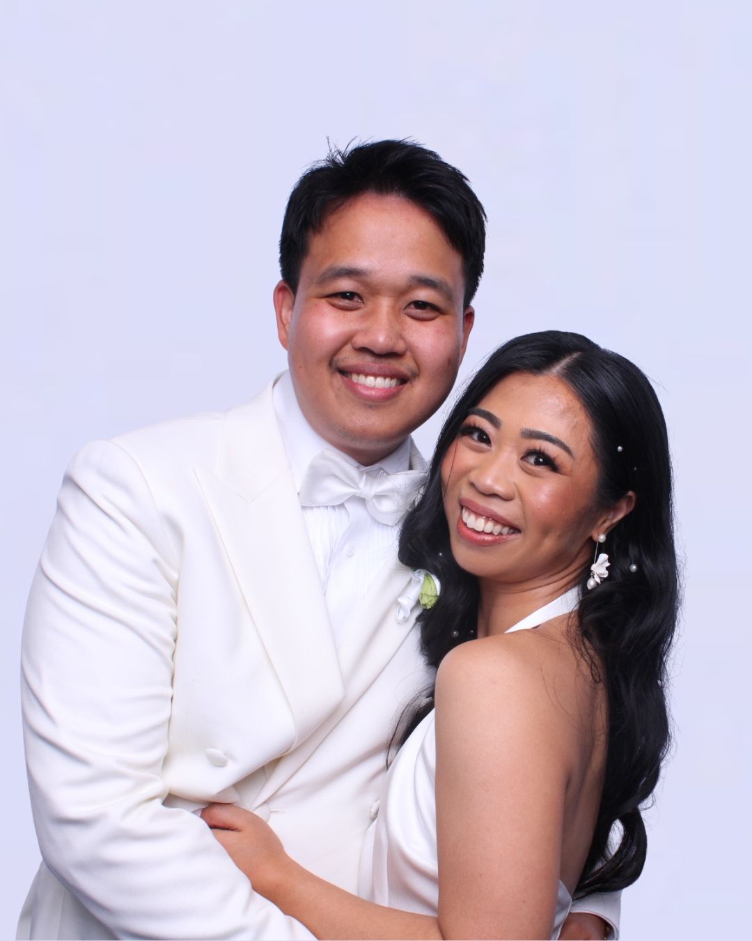 Smiling Asian couple embraces, wearing white wedding attire for Photo booth. 