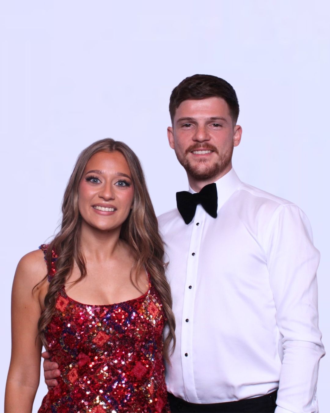 Woman in red sequin dress with man in tuxedo shirt and bow tie, both smiling wedding photo booth. 