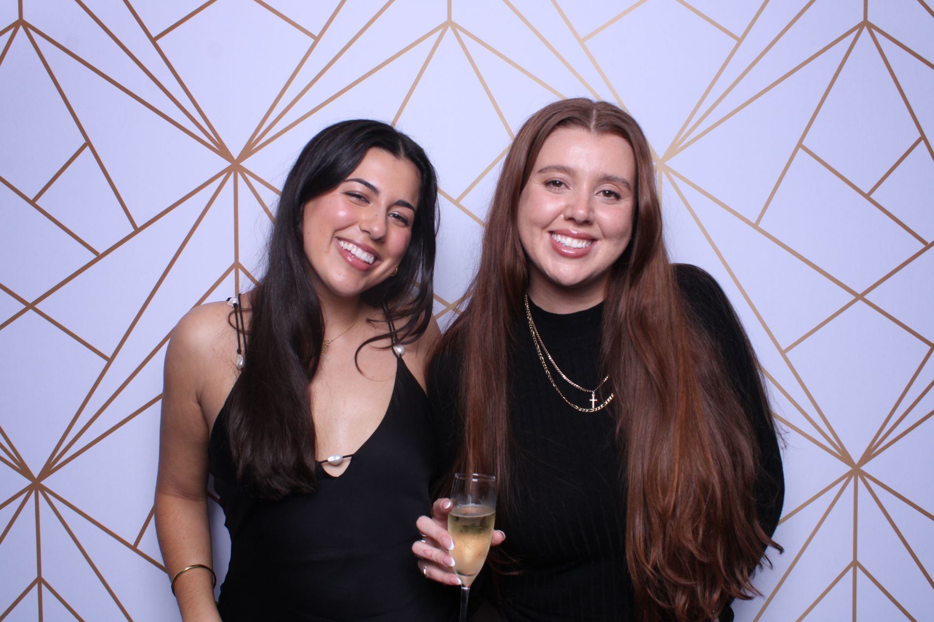 Two women smiling in a photo booth. One holds a champagne flute. Against a white and gold geometric backdrop.