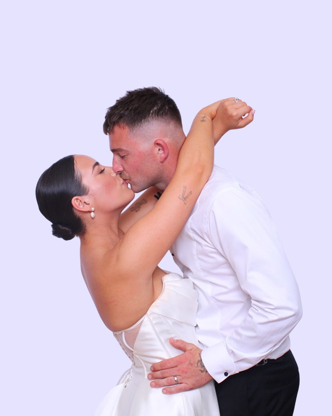 Bride and groom kissing in photo booth, white wedding dress, white shirt, leaning back, light blue background.