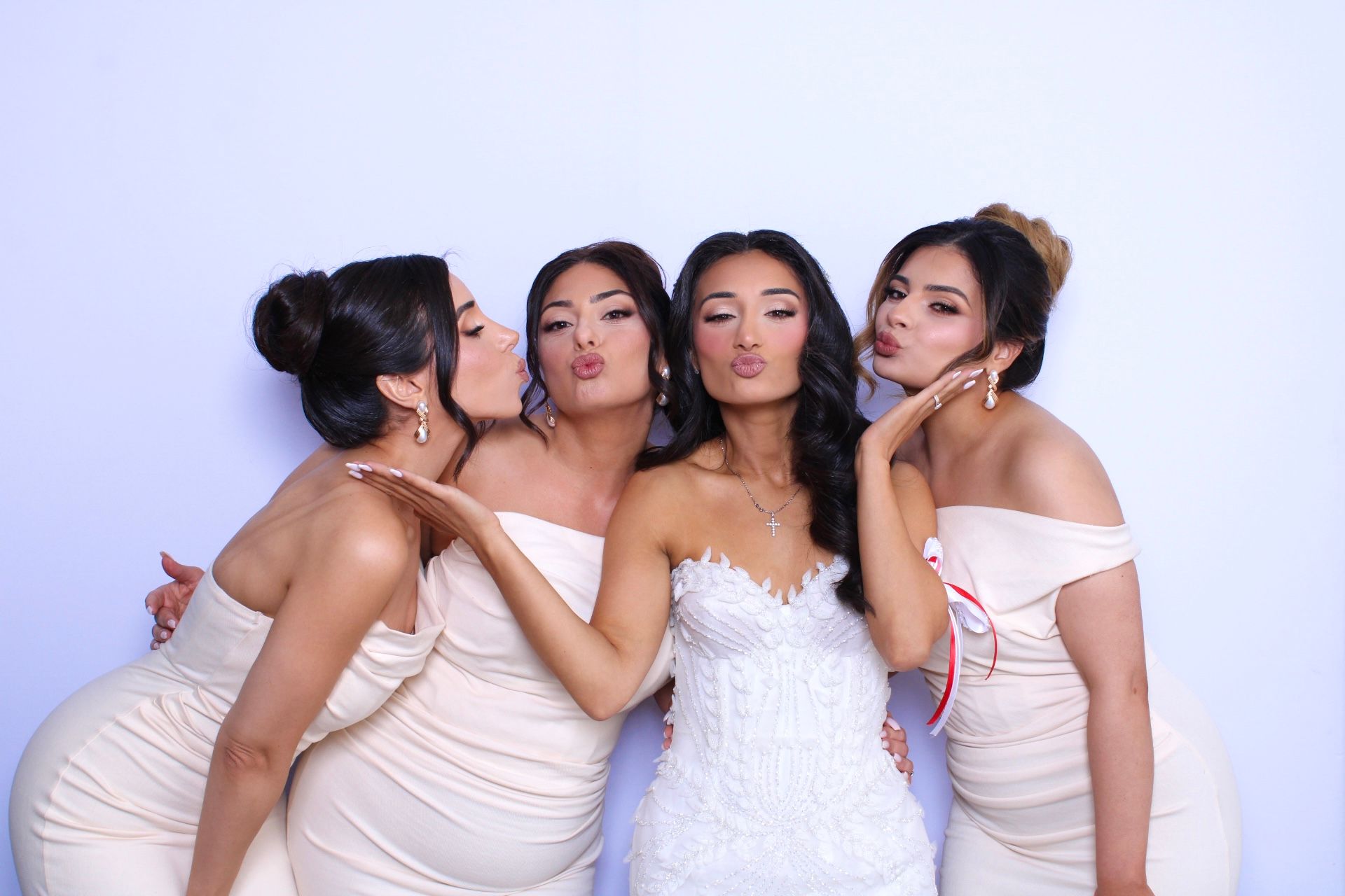 Bride in white dress with bridesmaids in beige, blowing kisses, white background in Photo Booth.