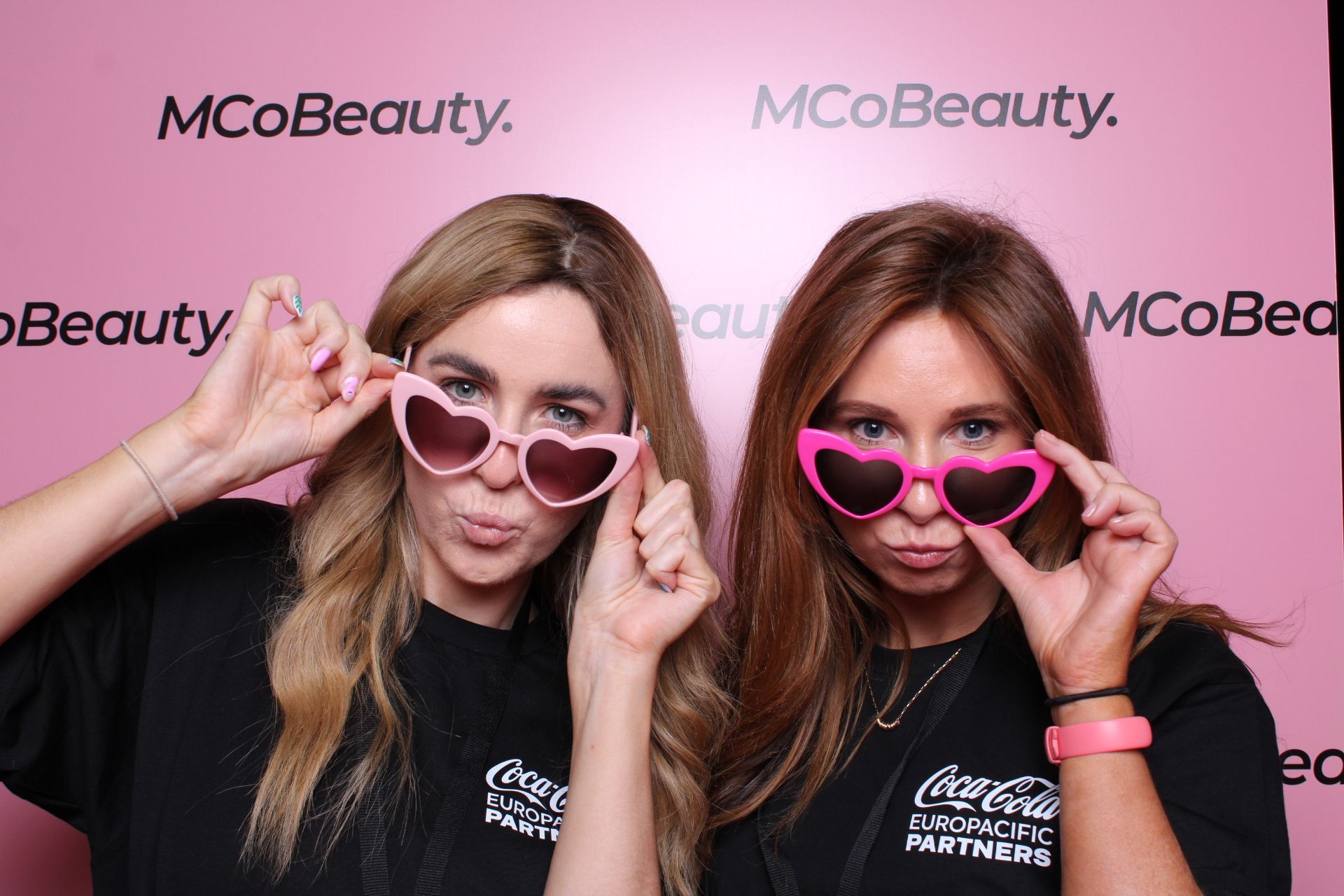 Two women wearing heart-shaped sunglasses, making playful faces in front of a pink MCoBeauty Makeup backdrop.
