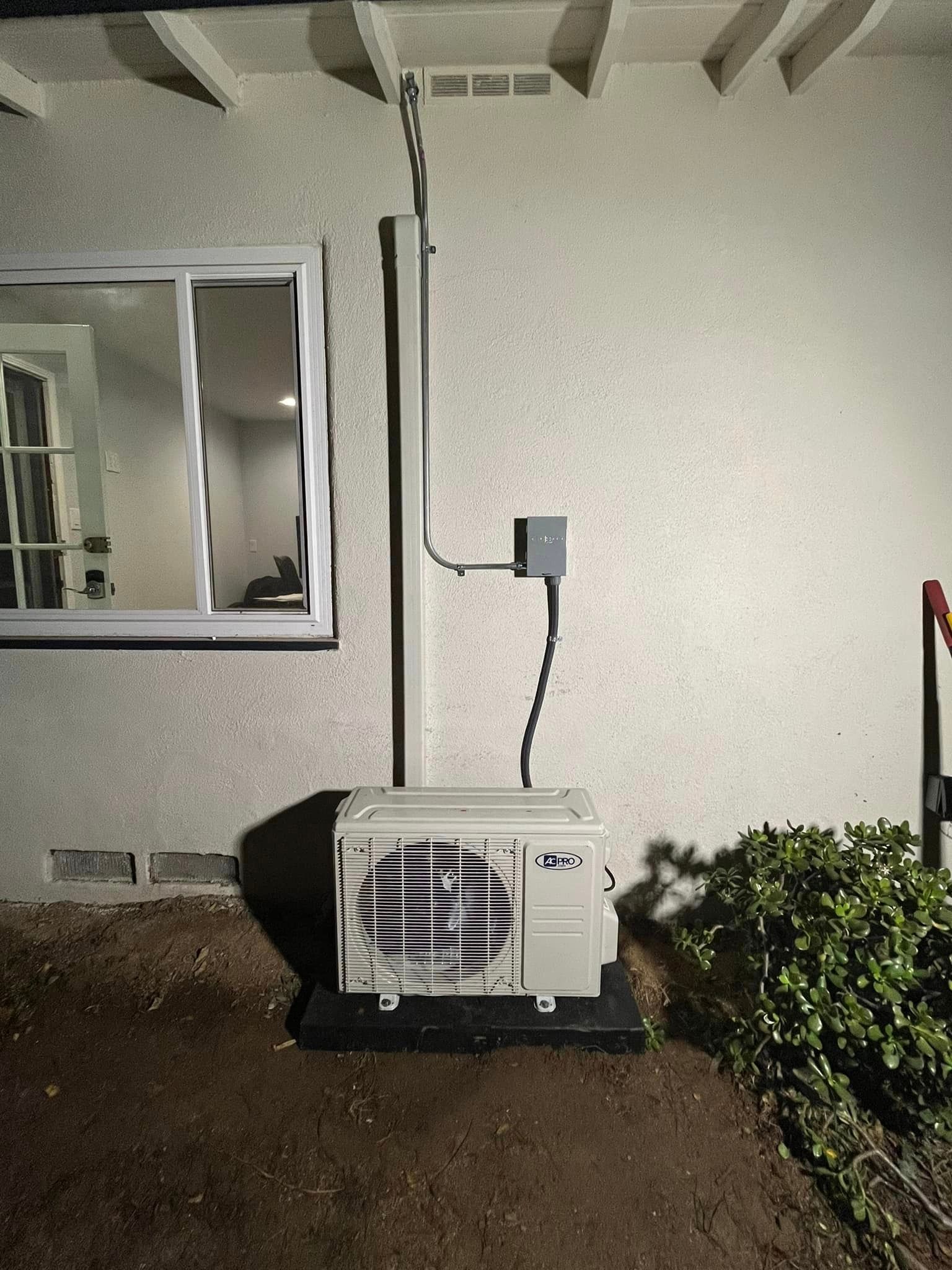 A white air conditioner is sitting on the side of a house next to a window.