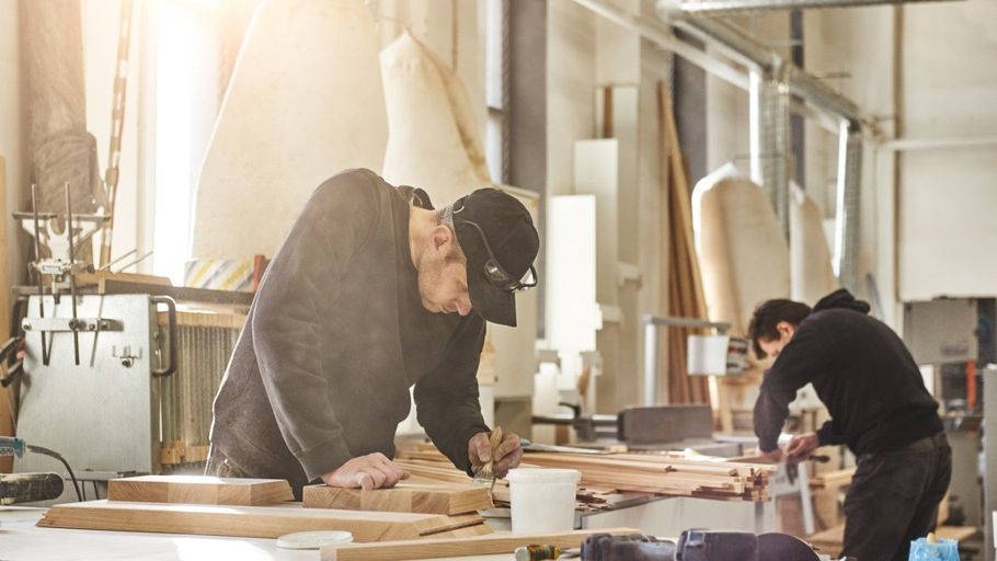 Two people work at a woodworking bench in a bright workshop, cutting and sanding wood.
