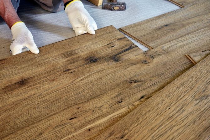 Gloved hands installing a wooden floorboard with a tool on a plank floor