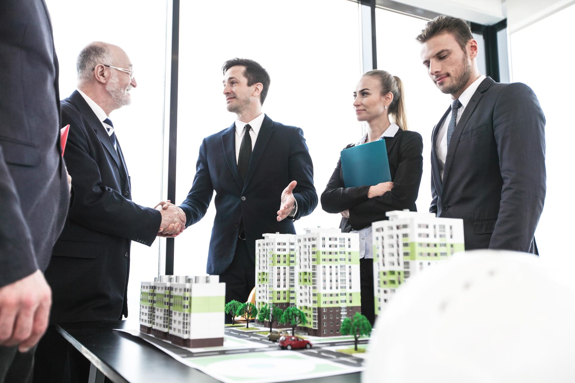Businesspeople shaking hands near architectural models, in an office setting.