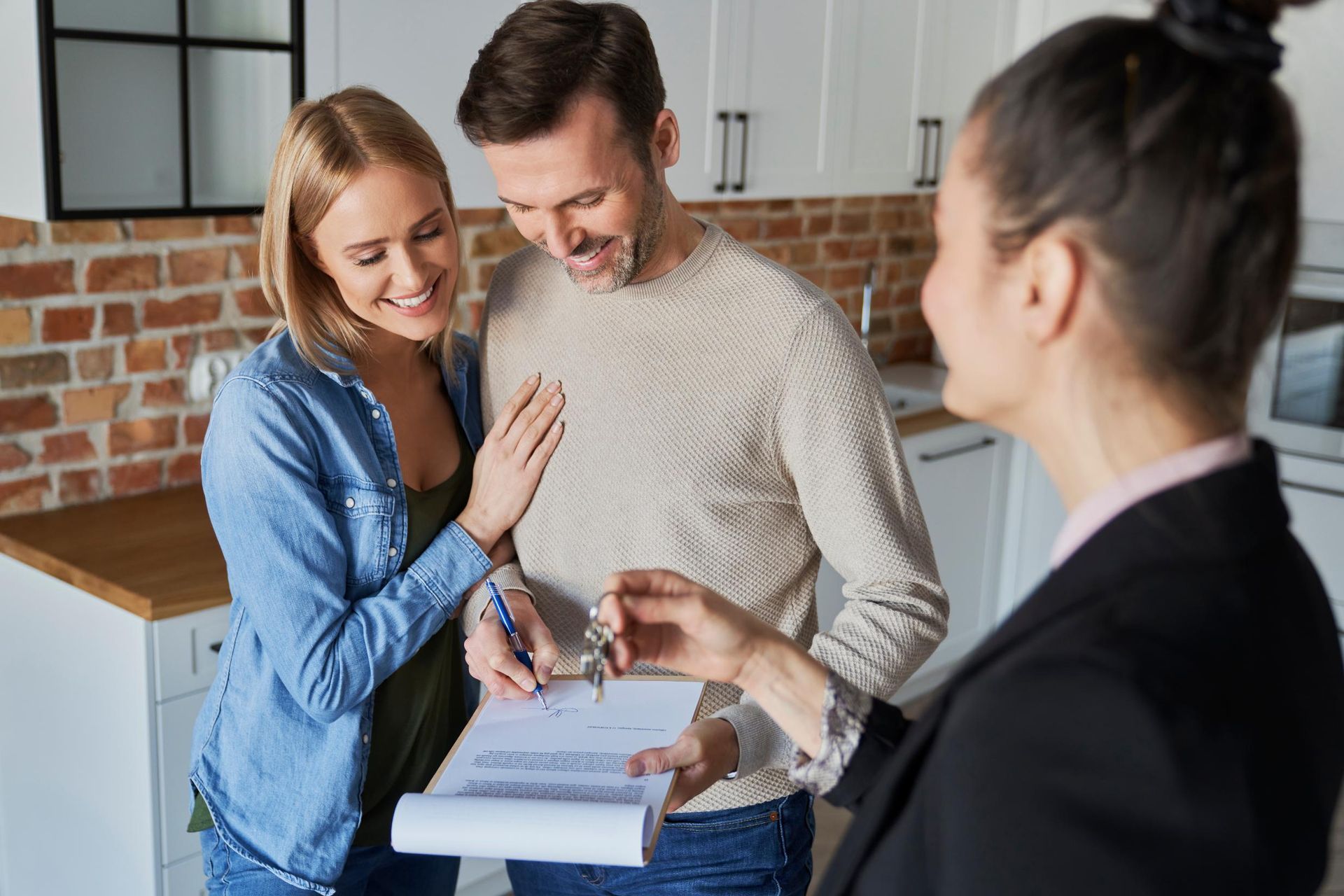 Couple signing a document in a kitchen, receiving keys from a realtor.