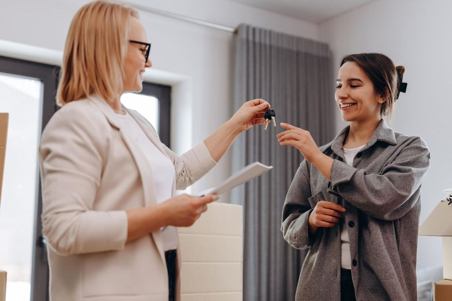 Woman handing keys to another woman; indoors, smiling, holding documents and moving boxes.