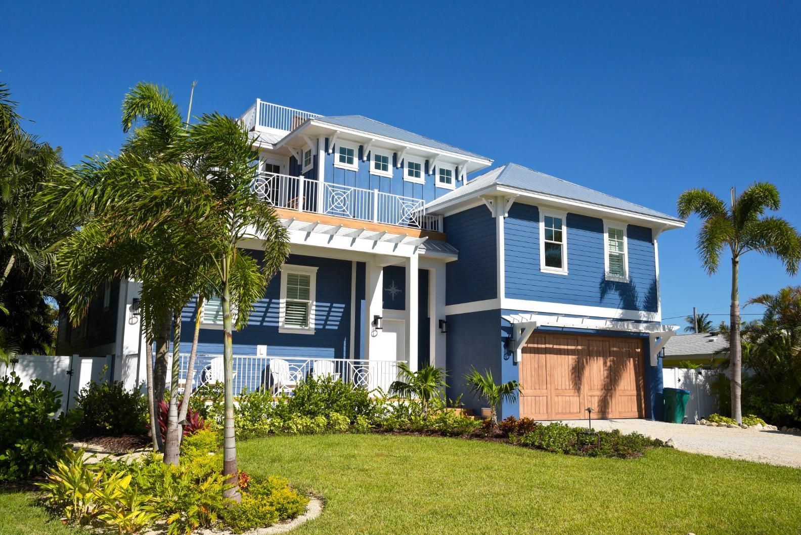 Blue beach house with tan garage door, lush lawn, and palm trees under a blue sky.