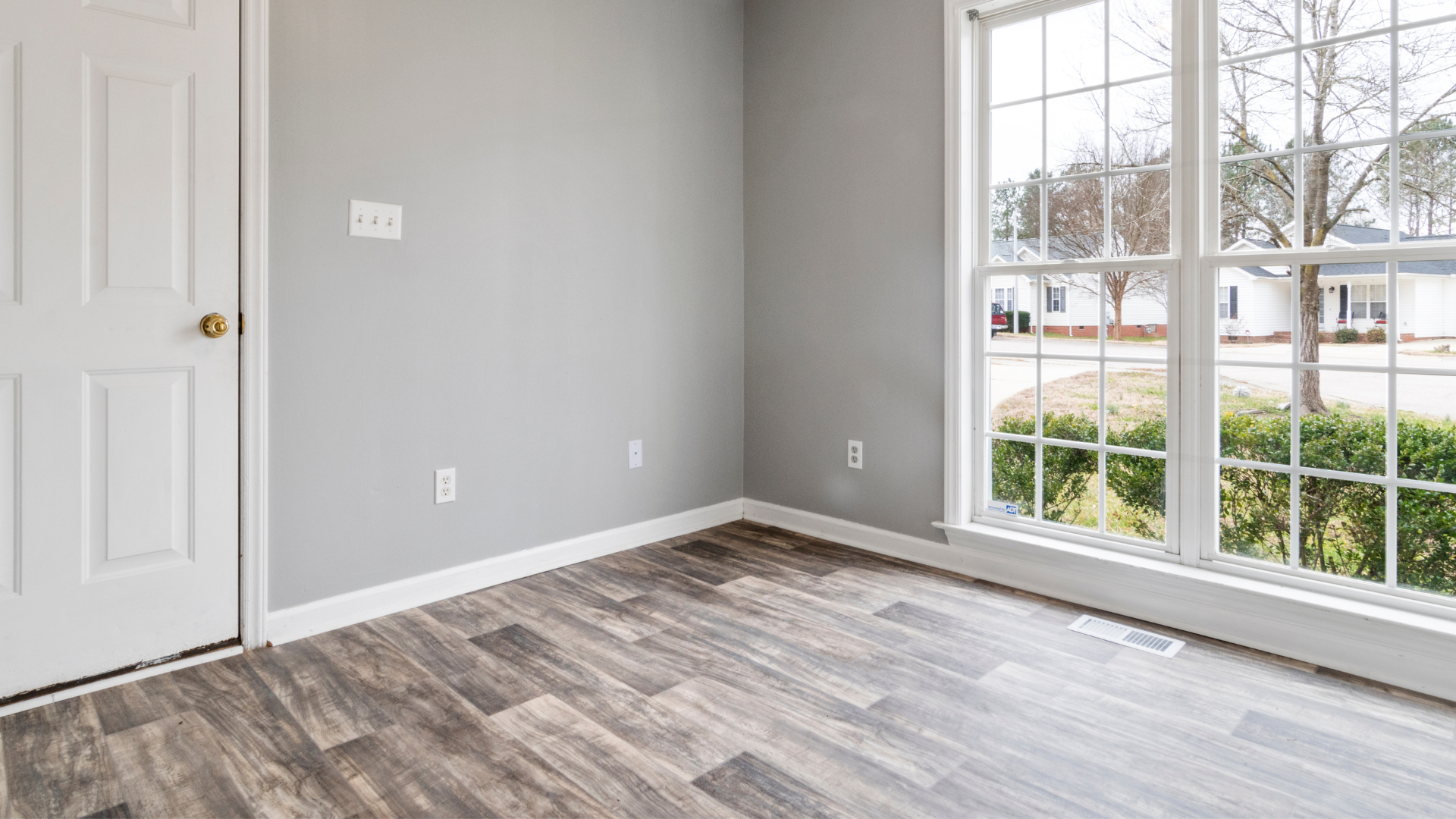 Empty room with gray walls, white trim, a door, and a large window overlooking a street.