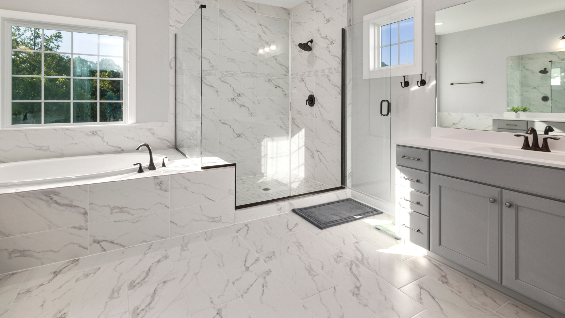 Spacious, modern bathroom with marble tile, a tub, glass shower, grey vanity, and natural light.